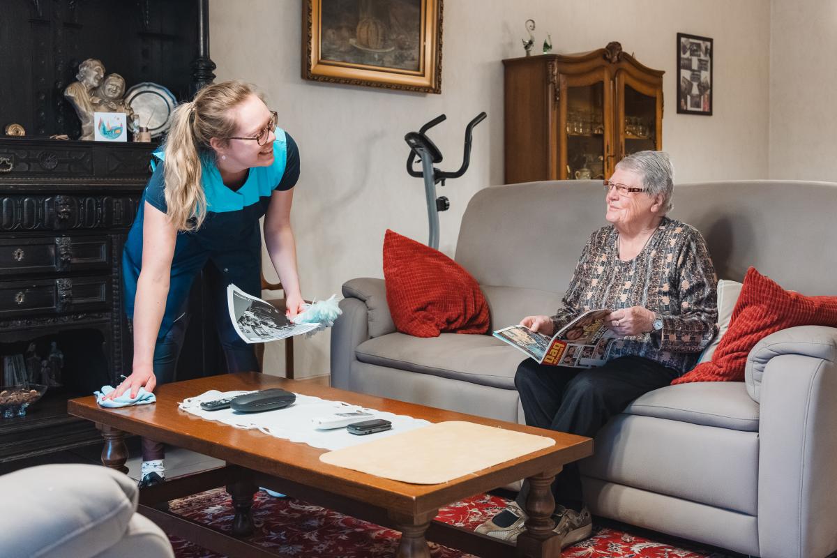 een poetshulp in de thuiszorg poets bij een oudere dame die in haar zetel zit